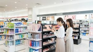 最新のコスメティックアイテムが並ぶ、日本の都市部にある明るく清潔な大型バラエティショップのコスメ売り場。棚にはチャコット製品も陳列されており、買い物を楽しむアジア人女性たちの姿が見える。